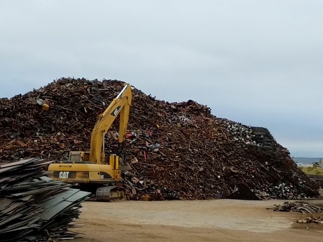 Heavy machinery operates near a large mound of scrap metal, highlighting a recycling or demolition site under a cloudy sky.
