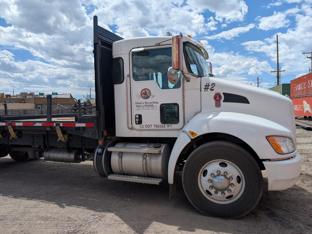 Flatbed truck from Steel & Recycling Iron & Metals parked on a gravel lot with an overcast sky in the background.