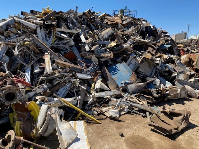 A large pile of various metal scraps and debris at a scrapyard under a clear blue sky, showcasing the recycling and salvage industry.